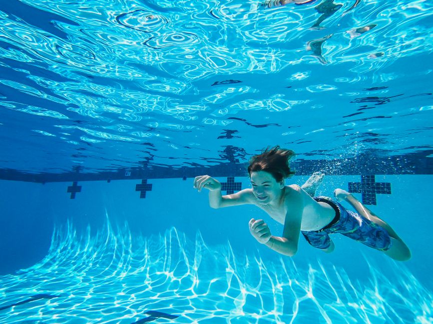 Rapaz nadando sorridente debaixo d'água em uma piscina, transmitindo diversão e descontração.