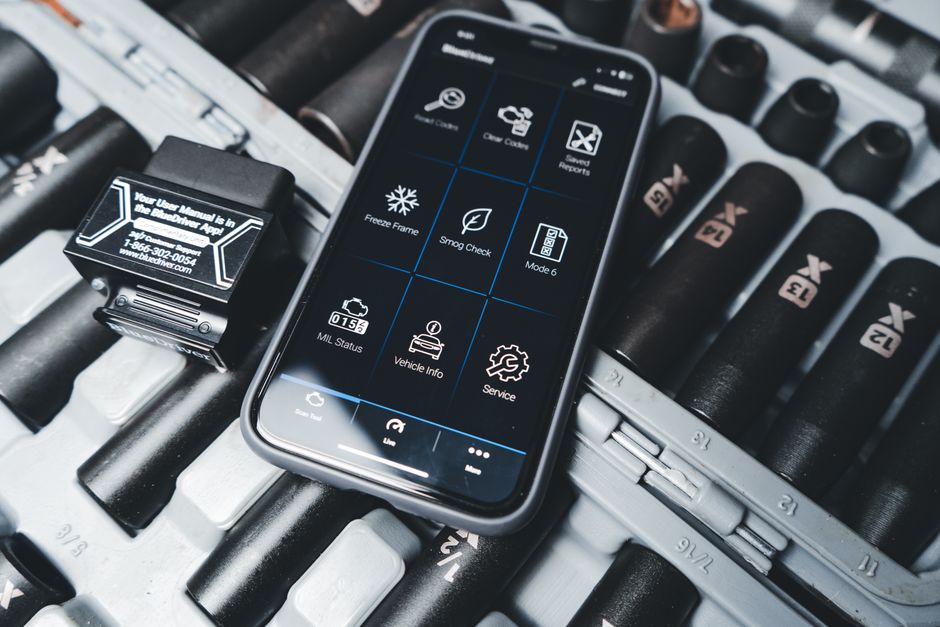 Imagem de um smartphone com aplicativo de controle de veículo, sobre uma caixa de ferramentas com chaves de roda, representando tecnologia aplicada à manutenção e gerenciamento de veículos.