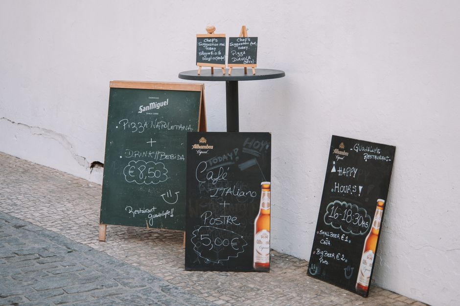 Quadro de avisos e promoções de um restaurante ao ar livre, com lousas pretas e de ar amador, exibindo menus e horários de funcionamento.