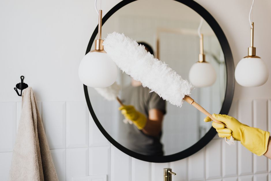 Imagem de uma pessoa limpando um espelho com um rolo de pintura, refletida em um espelho redondo decorativo, simbolizando manutenção e cuidado com o ambiente doméstico.