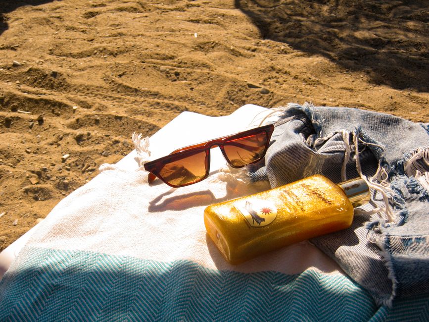 Óculos de sol, protetor solar e roupas de banho em uma praia, simbolizando lazer e cuidados com a saúde da pele ao ar livre.