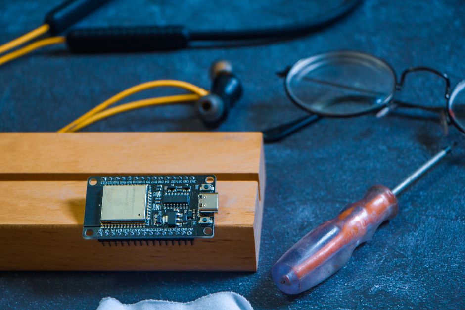 Imagem de uma placa de desenvolvimento eletrônica sobre um suporte de madeira, com óculos de leitura, fone de ouvido e uma chave de fenda ao redor, sugerindo um contexto de montagem ou programação de dispositivos tecnológicos.