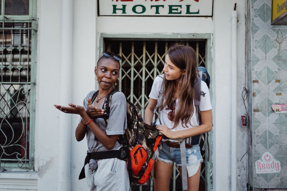 Duas jovens turistas conversando na rua em frente a um hotel, enquanto trocam dicas de viagem.