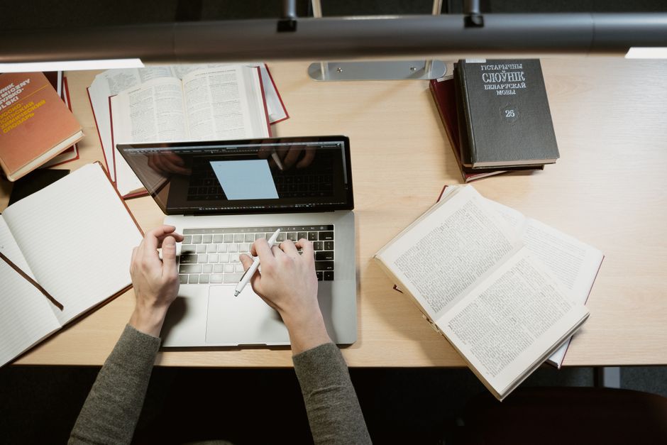 Pessoa estudando ou pesquisando com vários livros e um computador portátil em um ambiente de estudo ou trabalho acadêmico.