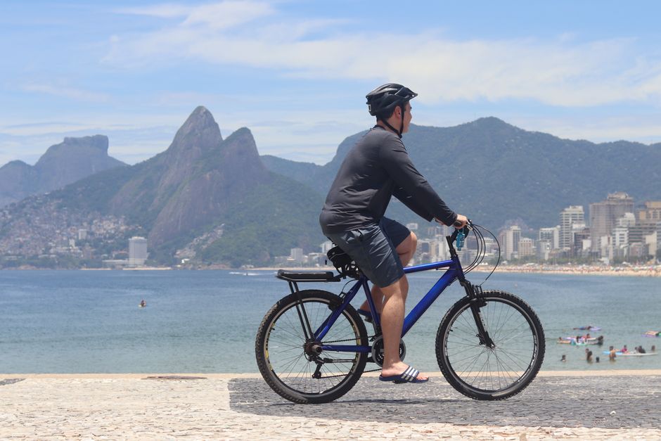 Homem pedalando de bicicleta à beira do mar com cenário de montanhas e cidade ao fundo, em uma manhã ensolarada.