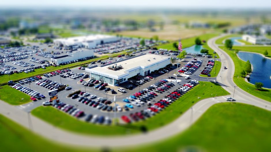 Imagem aérea de um grande centro de compras ou loja de varejo com estacionamento amplo, rodeado por áreas verdes e lagos, ilustrando o conceito de comércio e desenvolvimento comercial.