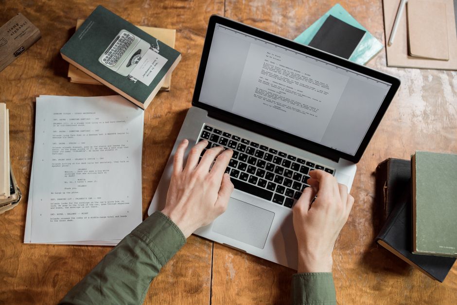 Pessoa trabalhando em computador com documentos e livros sobre escrita ou pesquisa acadêmica, em um ambiente de estudo ou escritório.