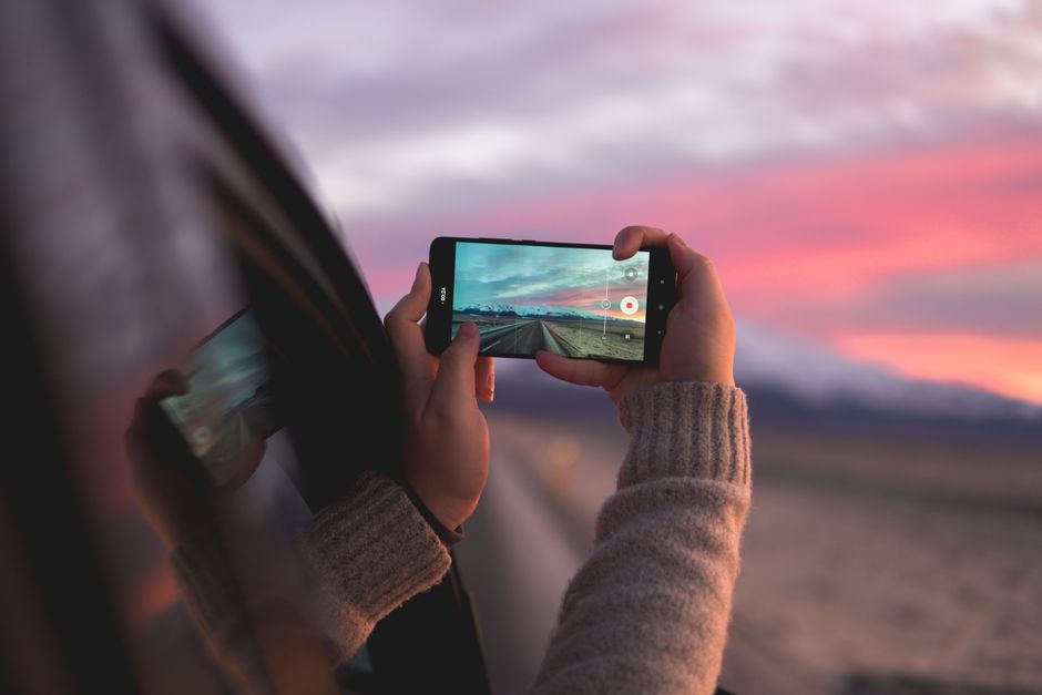 Pessoa gravando o pôr do sol com um smartphone enquanto está ao lado de um carro em movimento.