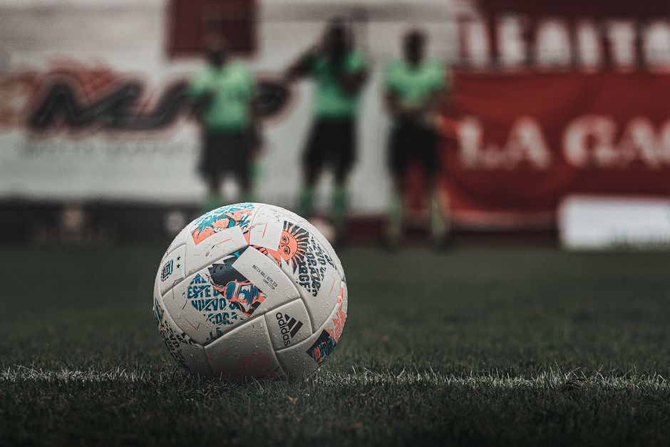 Imagem de uma bola de futebol em primeiro plano em um campo, com jogadores ao fundo, durante uma partida ou treino.