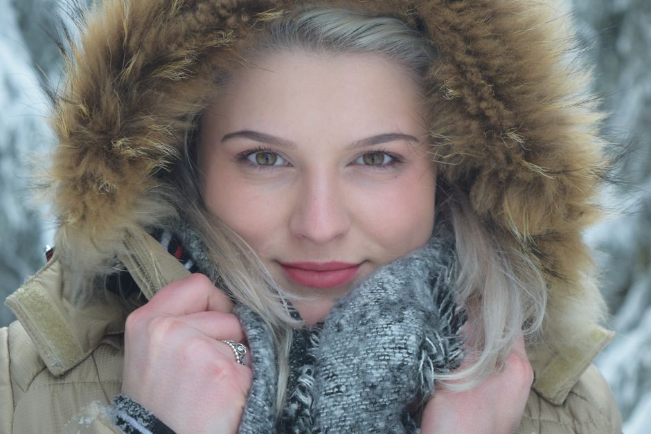 Mulher com visual invernal, usando casaco com capuz de pelúcia e cachecol, em ambiente de neve ao ar livre, transmitindo sensação de frio e aconchego.