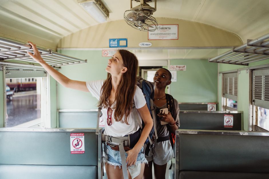 Duas jovens viajando de trem, uma delas ajustando o bagageiro enquanto a outra observa ao redor, em uma viagem de aventura.
