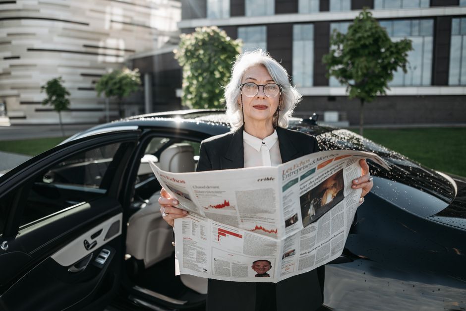 Mulher de aparência confiante lendo jornal financeiro ao lado de um carro, simbolizando negócios e análise de mercado.