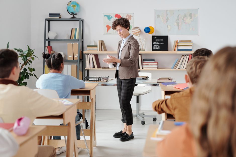 Professora dando aula para estudantes em uma sala de aula com mapas e livros ao fundo, demonstrando um ambiente de educação e aprendizado.