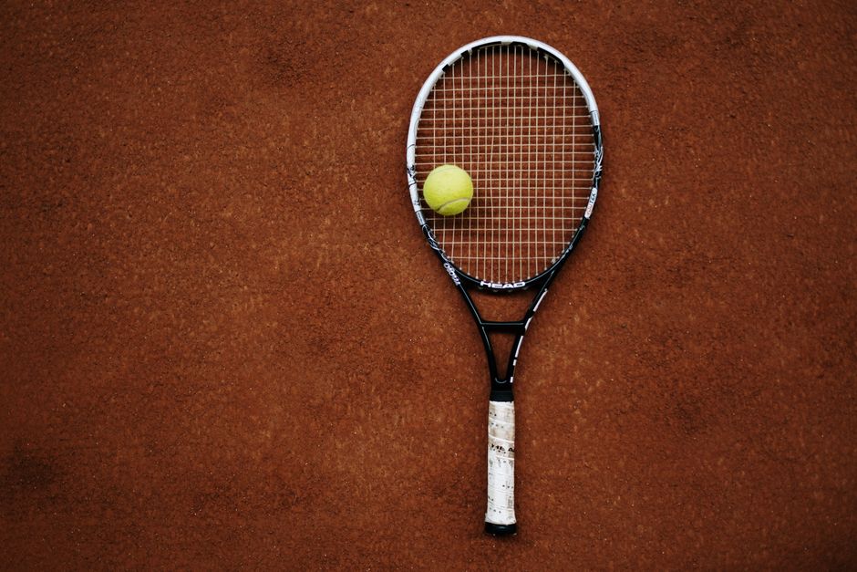 Raquete de tênis e uma bola dispostas sobre uma quadra de saibro, simbolizando o esporte de tênis.