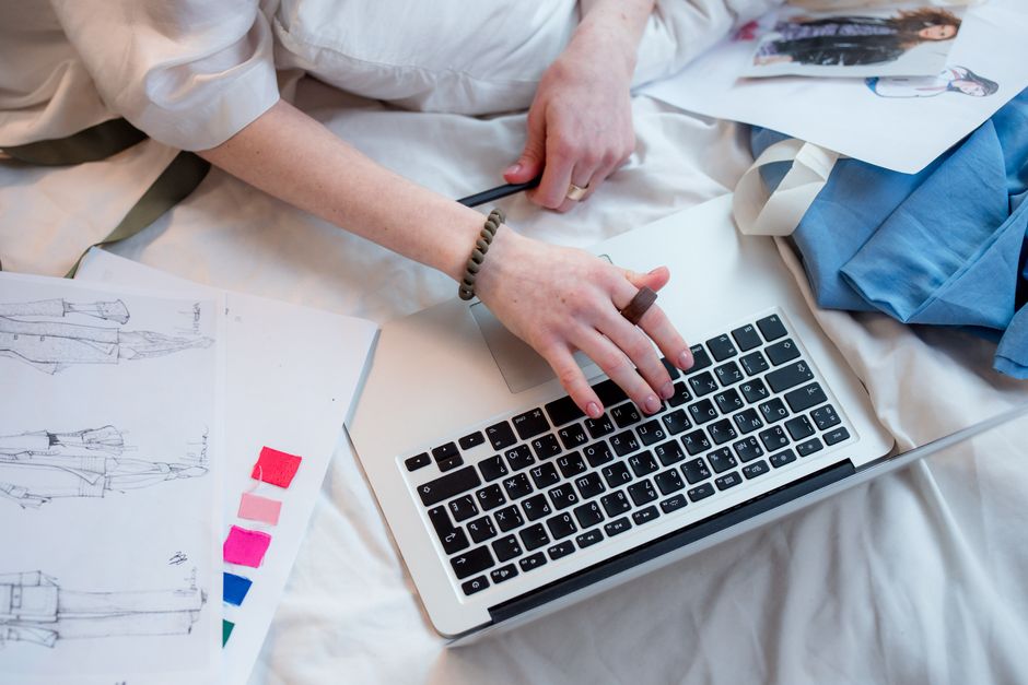 Pessoa trabalhando com computador e documentos, representando o planejamento criativo ou projeto de moda em um ambiente doméstico ou de estúdio.