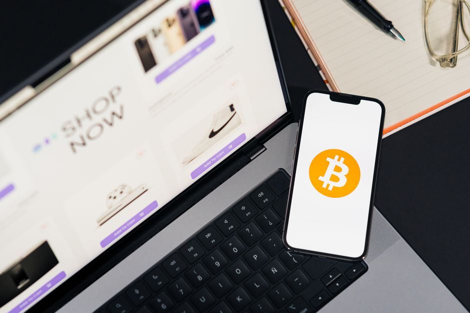 Imagem de um ambiente de trabalho com laptop exibindo uma loja online, celular com símbolo de Bitcoin, caneta, óculos e caderno, sugerindo temas relacionados a finanças digitais e investimentos.