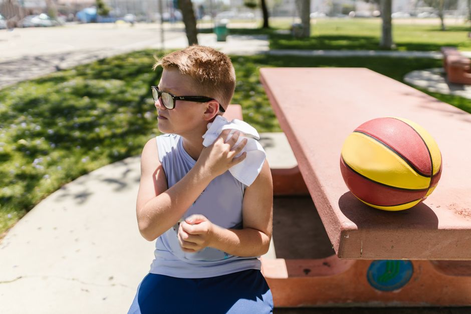 Rapaz jovem sentado em parque, segurando o ombro após partir-se ao se machucar enquanto brincava com uma bola de basquete ao lado, em um dia ensolarado.