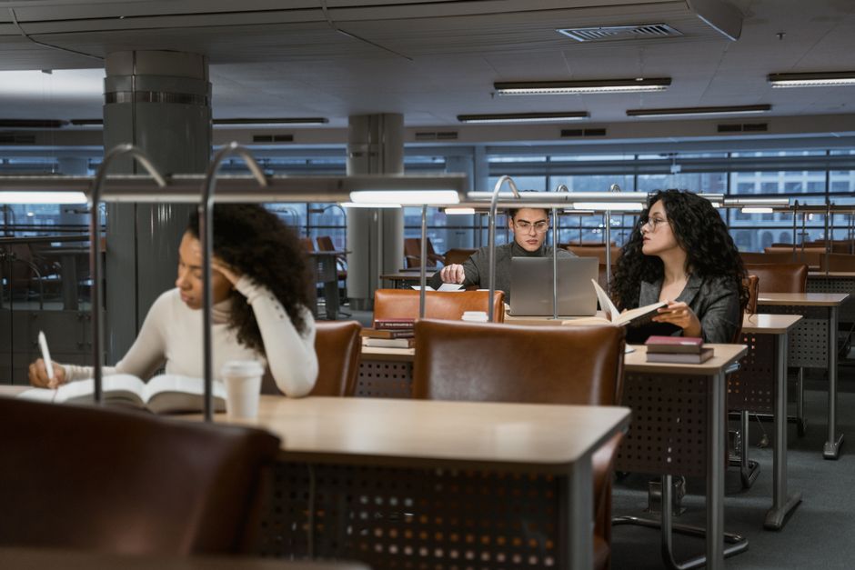 Três pessoas estudando ou trabalhando em um espaço de leitura ou pesquisa, usando computadores e livros em um ambiente corporativo ou acadêmico.
