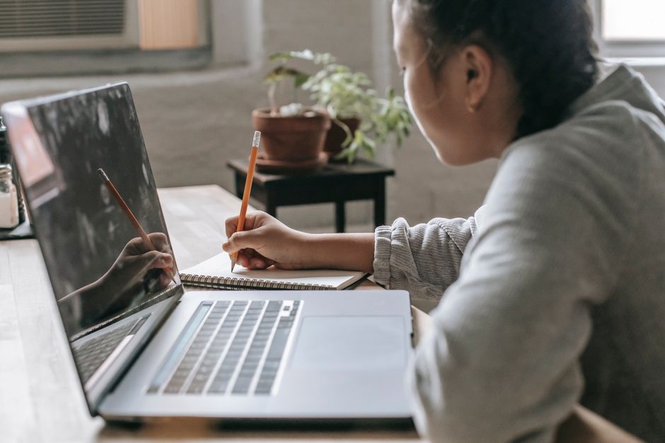 Pessoa estudando ou trabalhando remotamente com um laptop em um ambiente de casa, focada na tela e anotando em caderno.