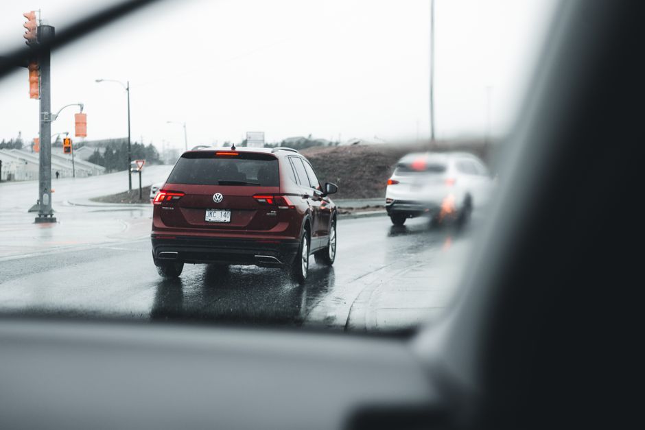 Imagem capturada de dentro de um carro mostrando uma rua movimentada com veículos em movimento sob clima nublado e úmido.