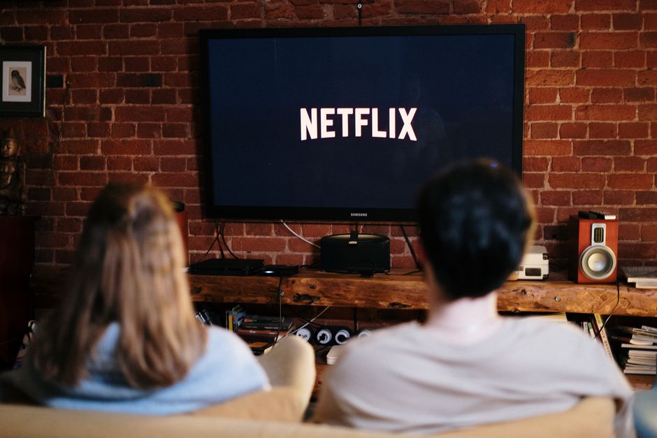 Dois adultos assistindo a uma série na Netflix em uma sala com parede de tijolos expostos.