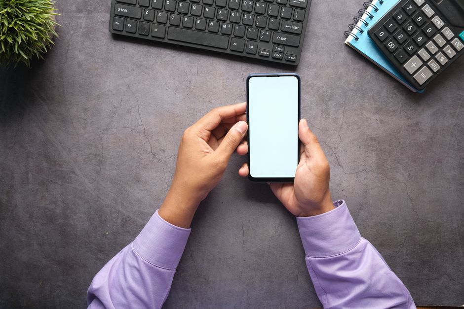 Pessoa segurando um smartphone em ambiente de trabalho com computador e documentos ao redor, representando produtividade e gestão digital.
