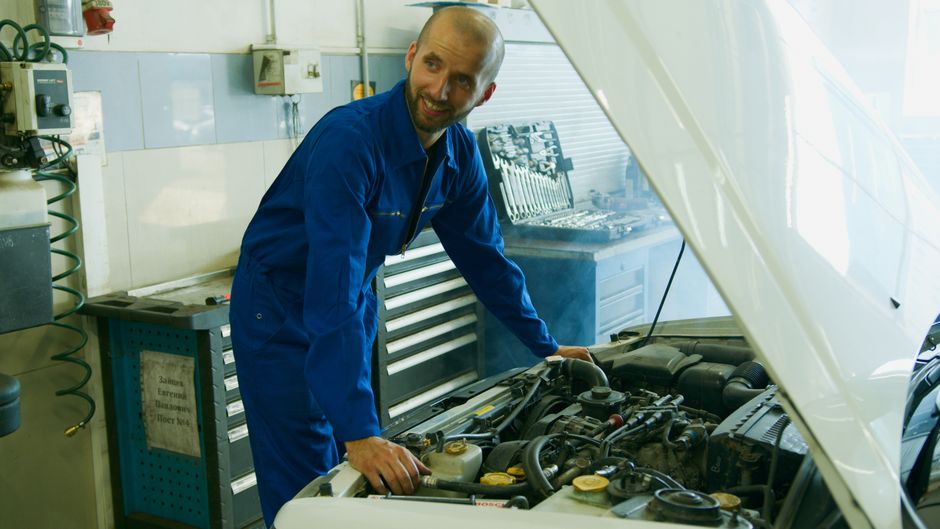 Imagem de um mecânico sorridente realizando manutenção em um carro no seu oficina, simbolizando trabalho técnico e reparos automotivos.