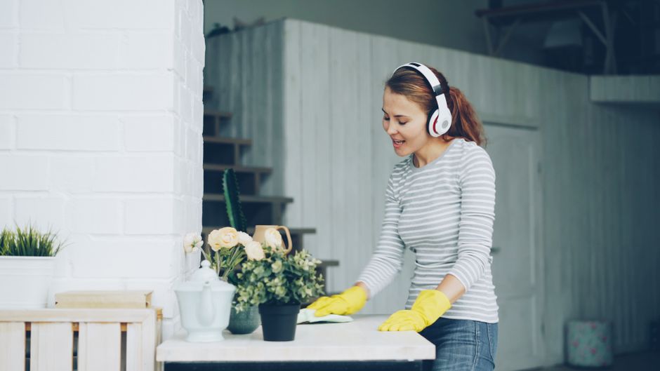 Mulher limpando plantas em ambiente interno, usando fone de ouvido e roupas casuais.