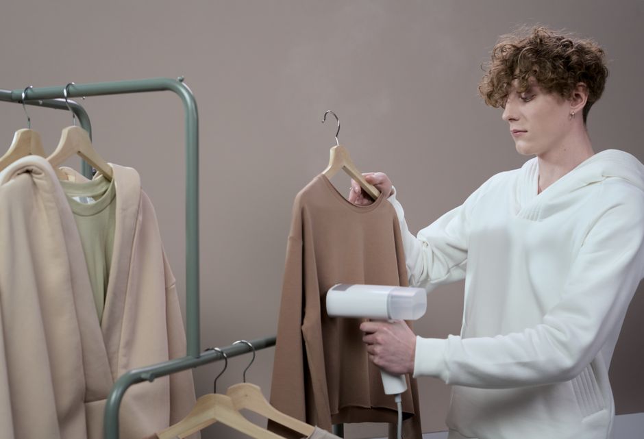 Pessoa passando roupa em uma peça de roupa de cor bege, utilizando um ferro de passar com painel digital, em ambiente interno neutro.