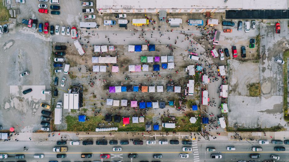 Vista aérea de um mercado ao ar livre com várias tendas coloridas e veículos ao redor, demonstrando um ambiente de comércio e interação social em uma cidade.
