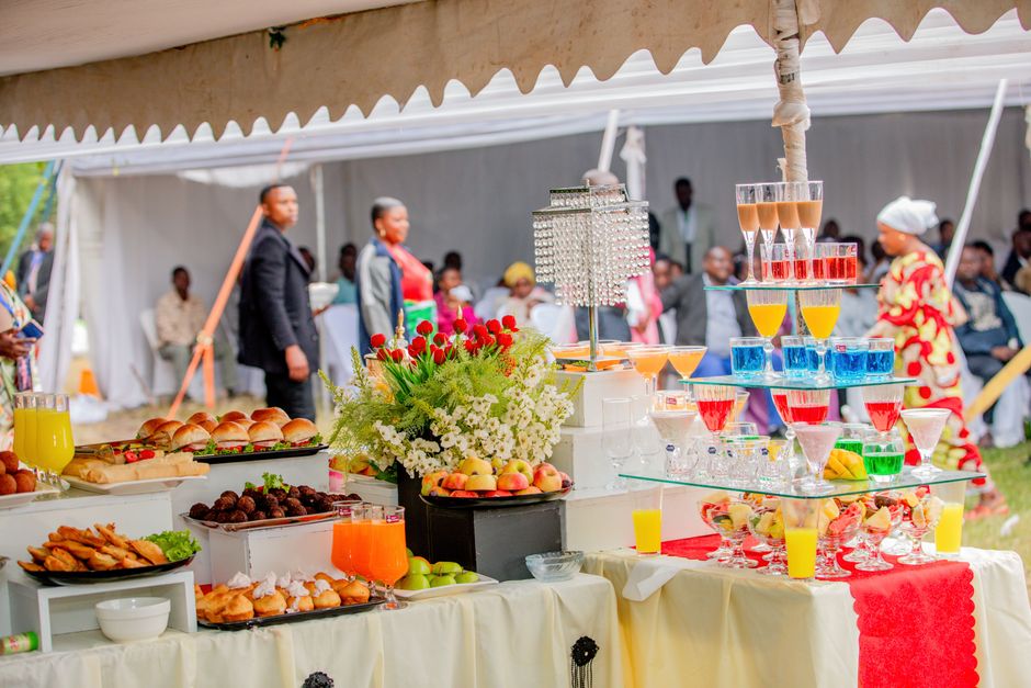 Mesa de festa ao ar livre decorada com frutas, doces e uma estrutura de bebidas coloridas, em um evento comemorativo com convidados e decoração festiva.