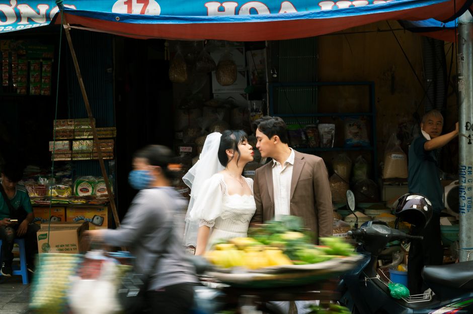 Casal de noivos posando em uma feira movimentada em um ambiente urbano, com vendedores e pedestres ao redor.