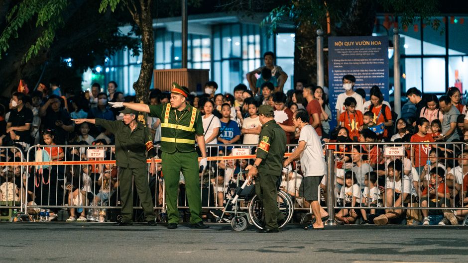 Imagem de uma organização de segurança em um evento público, com policiais orientando uma pessoa em cadeira de rodas diante de uma multidão em um ambiente externo à noite.