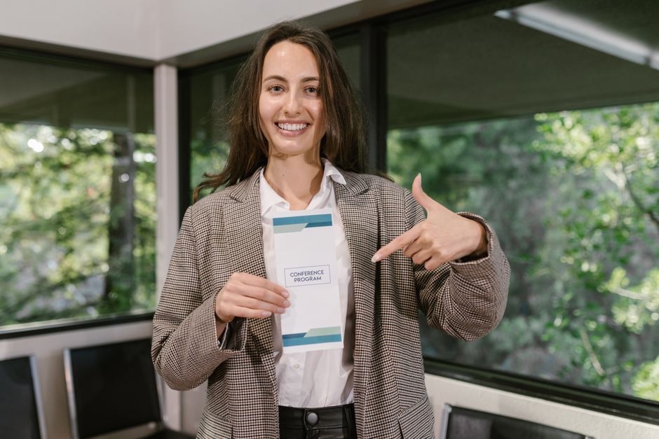 Mulher sorridente em ambiente corporativo segurando um programa de conferência, transmitindo entusiasmo e profissionalismo.