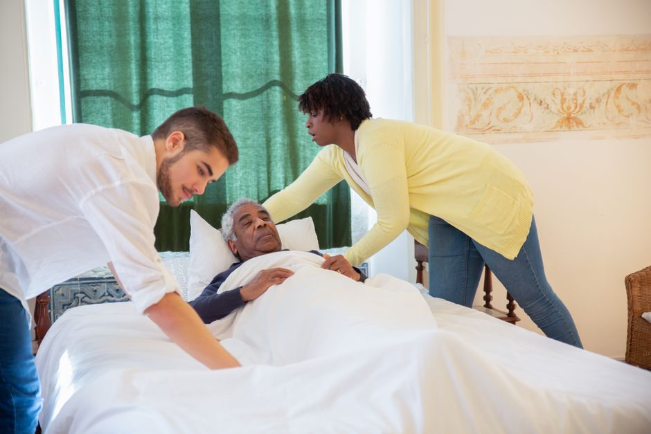 Imagem de duas pessoas cuidando de um idoso doente na cama, simbolizando apoio e cuidado na área da saúde.