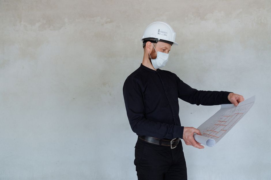 Engenheiro ou arquiteto avaliando projetos ou planos em um ambiente de obra, usando capacete e máscara de proteção.