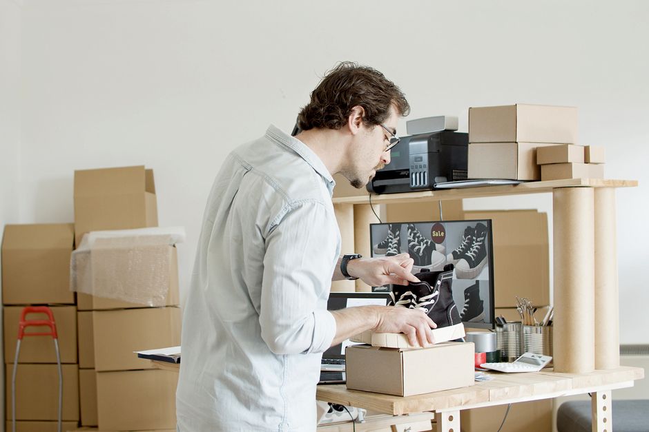 Homem organizando e embalando itens em um ambiente de trabalho com caixas de papelão e equipamentos de escritório, sugerindo processo de embalagem ou logística.