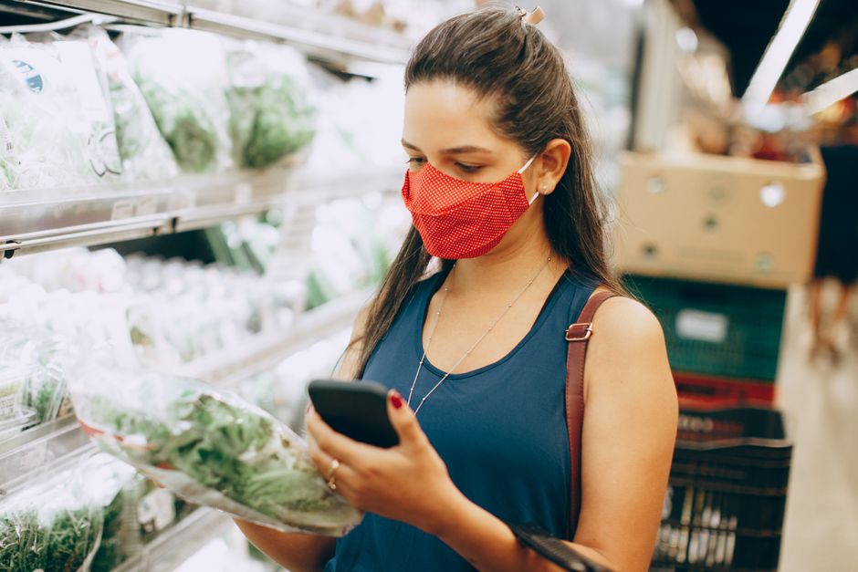 Mulher usando máscara facial verifica produtos frescos em supermercado, refletindo sobre compras conscientes e cuidados de saúde.