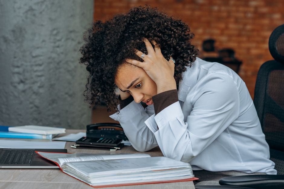 Mulher estressada lidando com várias tarefas e documentos em um ambiente de trabalho, refletindo sobre dificuldades no ambiente profissional ou acadêmico.