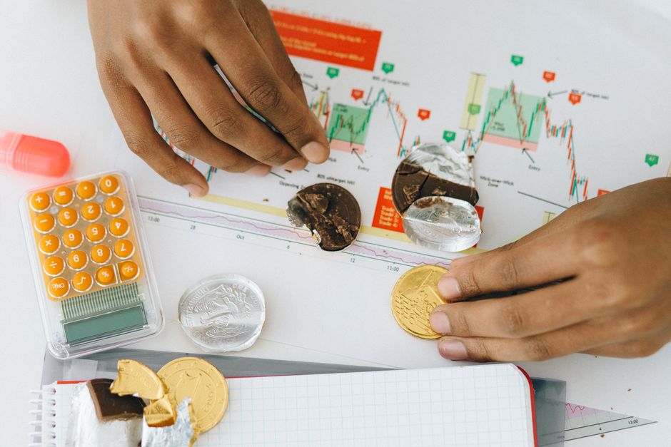 Pessoa analisando gráficos financeiros ao lado de chocolates e moedas, destacando a relação entre gerenciamento financeiro e consumo pessoal.