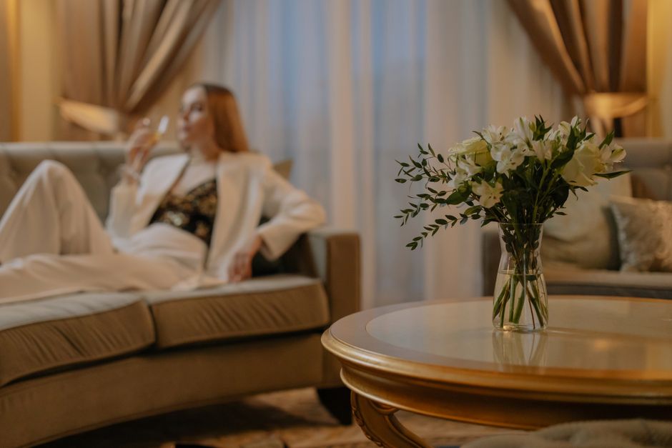 Mulher relaxando sozinha em um ambiente sofisticado, com destaque para uma mesa com vaso de flores no primeiro plano, transmitindo elegância e tranquilidade.
