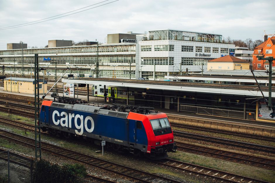 Imagem de uma estação de trem com um locomotivo de carga em primeiro plano e diversos prédios ao fundo, representando transporte e logística urbana.