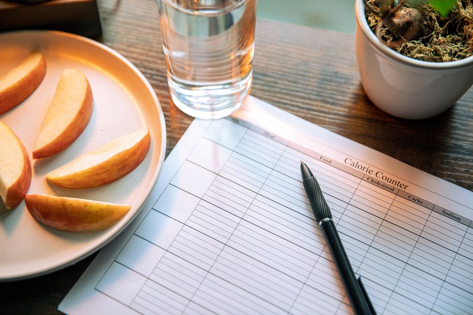 Imagem de uma rotina de planejamento de saúde e bem-estar, com uma planilha de controle de alimentação ao lado de uma taça de água, frutas e uma planta, ideal para um artigo sobre hábitos saudáveis.
