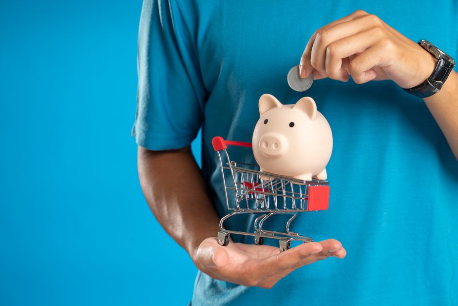 Pessoa colocando moeda em porquinho de porcelana dentro de um carrinho de supermercado para ilustrar temas de economia doméstica ou poupança.