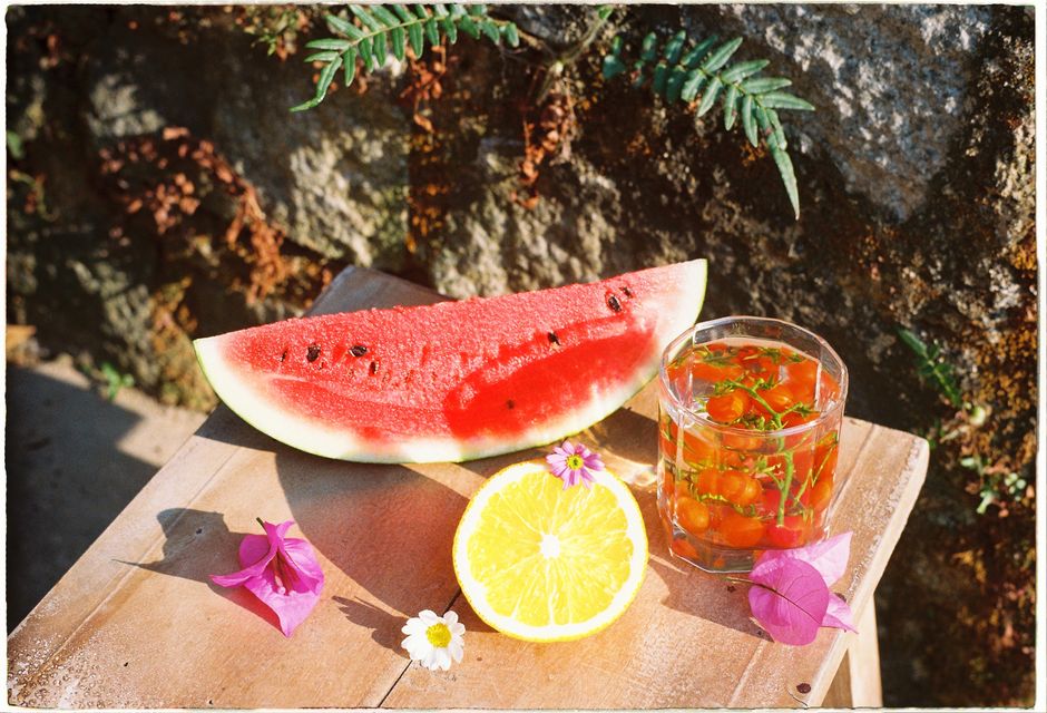 Foto de uma fatia de melancia, um copo de água com frutas e algumas flores, transmitindo uma sensação de verão e bem-estar ao ar livre.