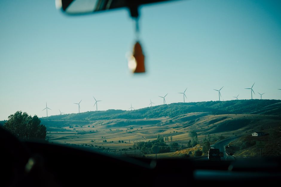 Imagem de uma paisagem rural vista do interior de um carro, com turbinas eólicas ao fundo, simbolizando energias renováveis e sustentabilidade.