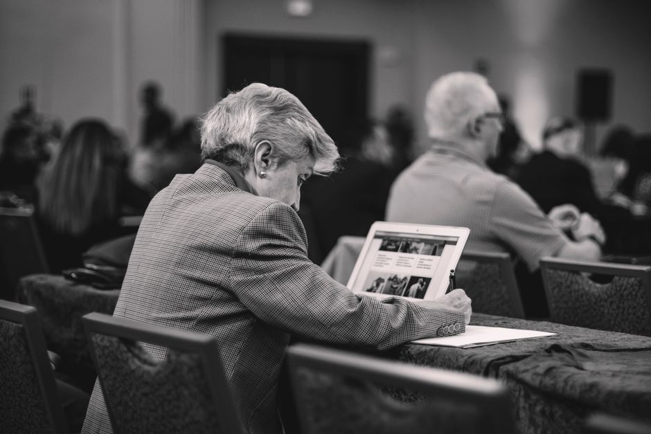 Pessoa idosa assistindo a uma apresentação ou reunião em um congresso ou seminário profissional, com atenção ao conteúdo exibido em um dispositivo digital.