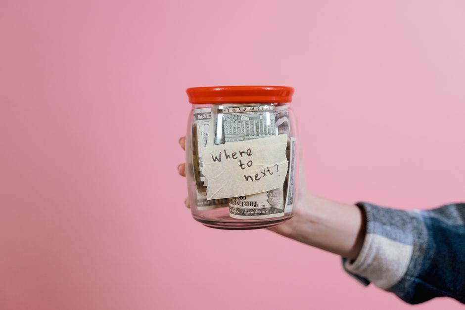 Pessoa segurando um pote de vidro cheio de dinheiro com um bilhete que diz 'Where to next?', simbolizando planejamento financeiro ou escolhas de investimentos.