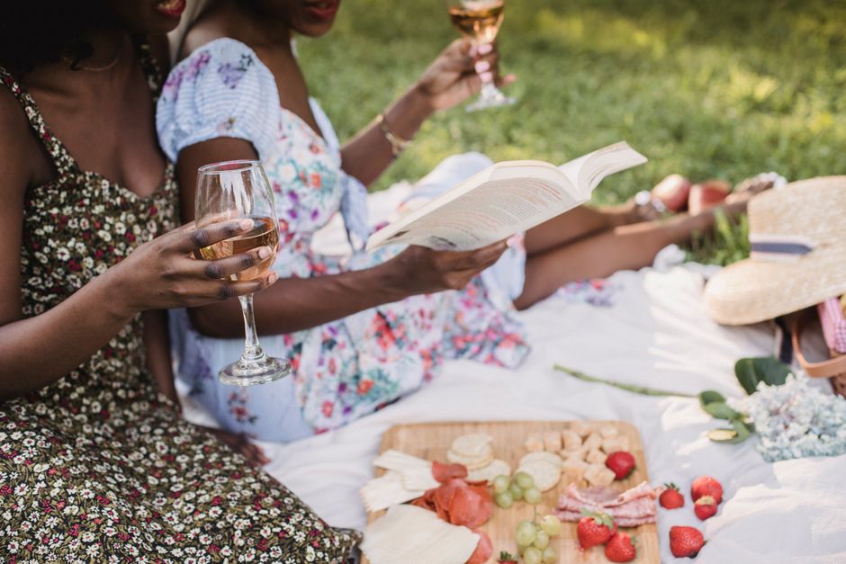 Mulheres desfrutando de um piquenique ao ar livre, com bebidas e leitura, em um ambiente de lazer e socialização.