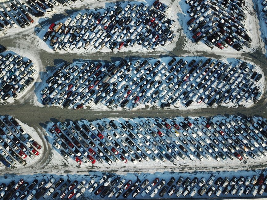 Imagem aérea de um grande estacionamento de carros cobertos de neve, destacando a quantidade e organização dos veículos.